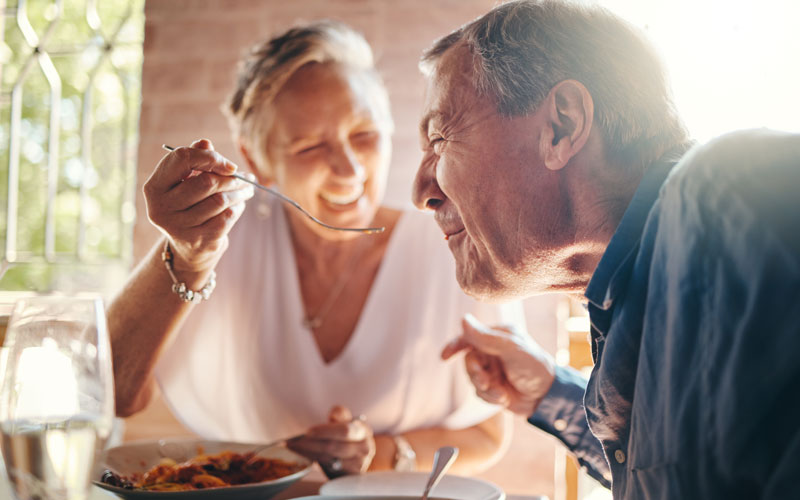 seniors-dining Elderly woman feeding elderly man with a spoon in a sunlit dining setting, both smiling warmly.