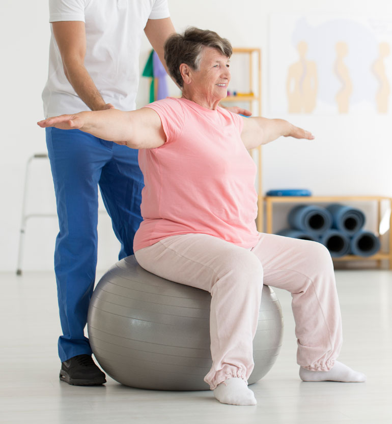 senior-therapy-session A senior woman sits on an exercise ball, guided by a therapist during a physical therapy session