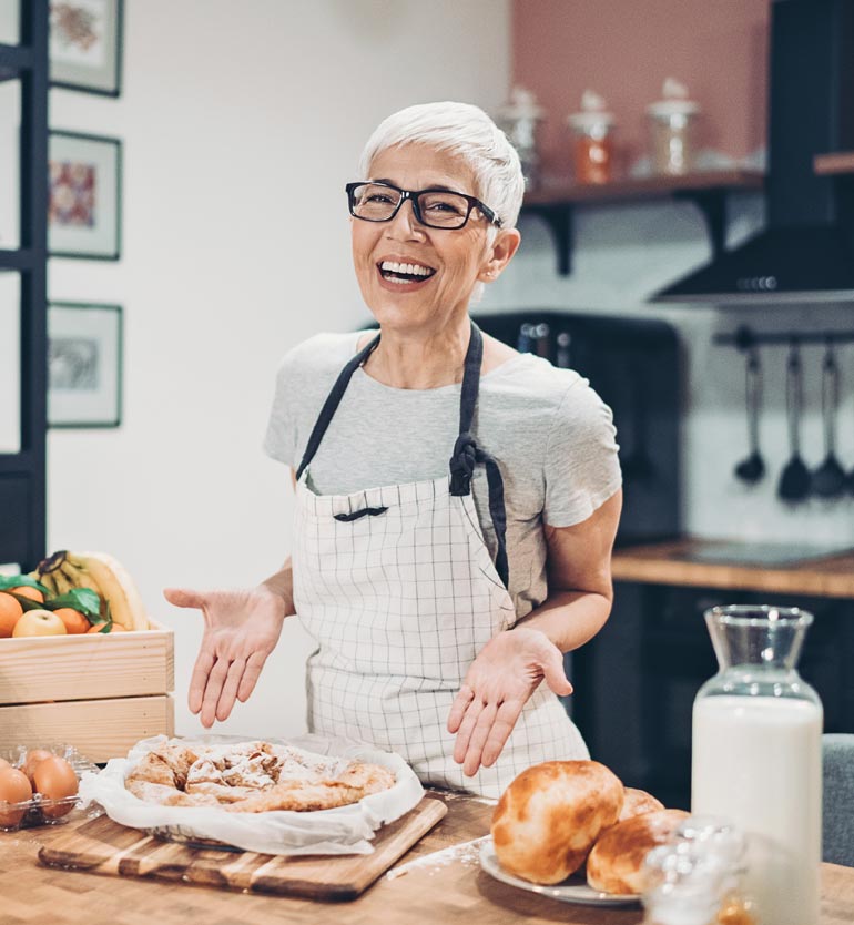 senior-baking Senior woman with glasses and apron baking in a kitchen, showing off a freshly baked dish.