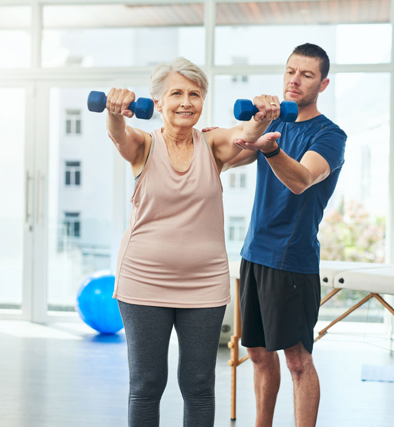 senior-lifting Senior woman lifting weights with a trainer assisting in a bright, modern gym setting.