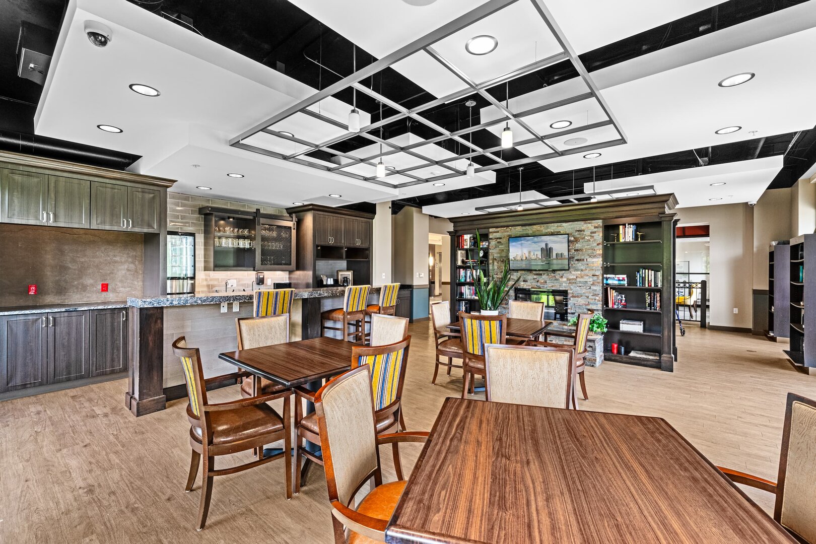 community-lounge-area-senior-living Spacious lounge with wooden tables, chairs, bookcase, and a television in senior living community.